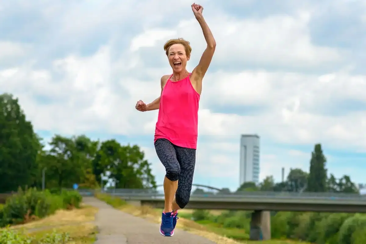 A joyful middle-aged woman in a pink tank top leaping with a raised fist on a riverside walking path